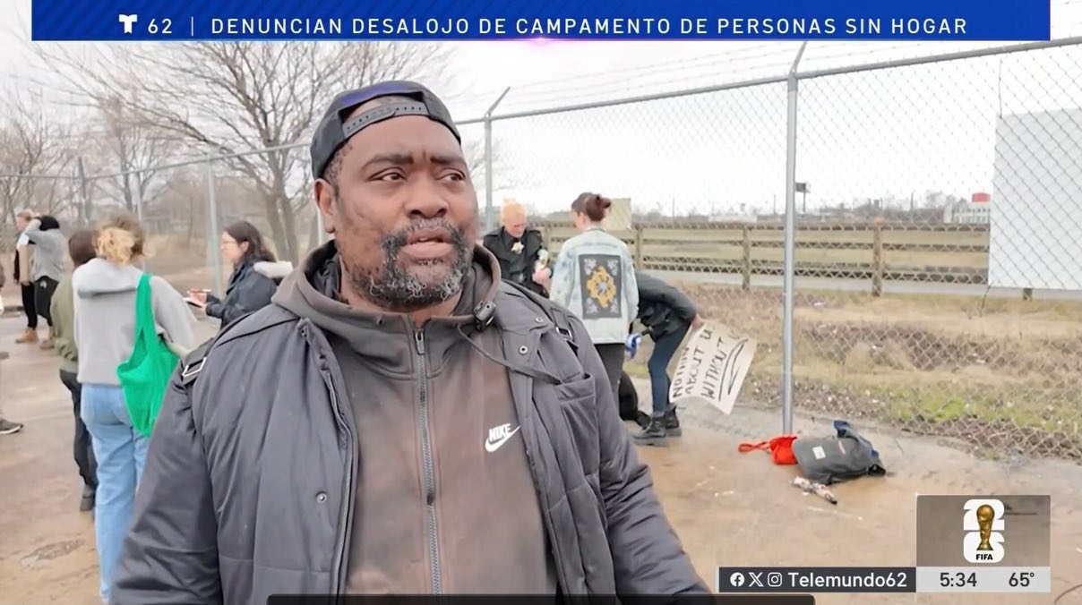 Man in hoodie standing in front of people holding signs by homeless encampment